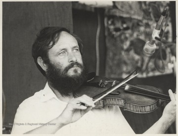 Franklin George is pictured playing a fiddle behind a microphone. This photograph is from a series of photos from Appalachian Folk Music Festivals, including the Ivydale and John Henry festivals. 