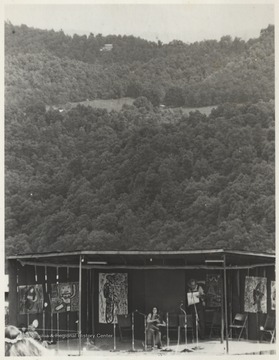 An unidentified woman performs on a stage positioned below a tree-covered hillside during a festival at Pipestem.  This photograph is part of a series of photographs showing Appalachian Folk Music Festivals, including the Ivydale and John Henry Festivals.