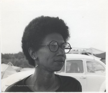 Beverly Cole is pictured at a campsite, with parked cars and pitched tents behind her. This photograph comes from a series of photos from Appalachian Folk Music Festivals, including the Ivydale and John Henry festivals. 