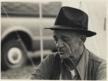 A man wearing a hat is pictured sitting outside of a tent. This photograph comes from a series of photos from Appalachian Folk Music Festivals, including the Ivydale and John Henry festivals. 