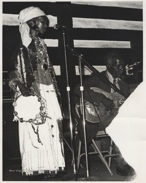 A woman, possible Della Taylor, with a tambourine watches as her associate, the Reverend Pearly Brown, plays the guitar on stage. This photograph comes from a series of photos from Appalachian Folk Music Festivals, including the Ivydale and John Henry festivals. 