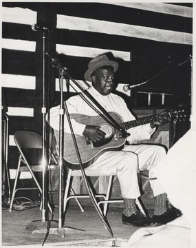 John Jackson plays a guitar and sings while seated behind a microphone. This photograph comes from a series of photos from Appalachian Folk Music Festivals, including the Ivydale and John Henry festivals. 