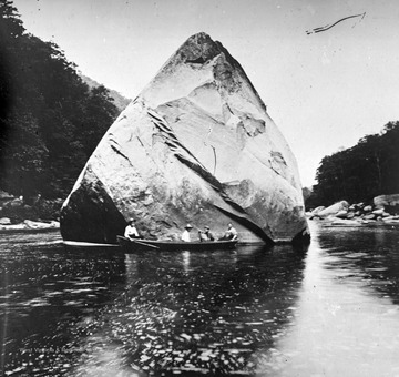 Four men sit in a canoe beside the large boulder in the middle of the river. Subjects unidentified. 