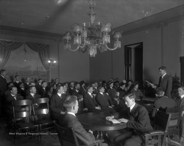 A group of students are gathered in a room. Many are listening to a man who is standing and reading from a sheet of paper. Subjects unidentified. 