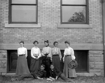 Five female students stand while two male students pose in the grass for a group portrait near a building that is likely the Central Public School in Morgantown, W. Va.  The Central Public School was built in 1898-1899 and was opened in September 1899 to grade and high school students.