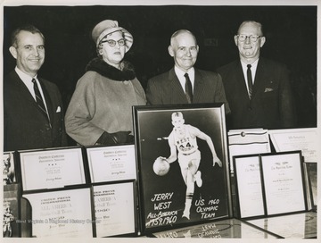 Mr. and Mrs. West are pictured in the center in between two unidentified gentlemen. The four are standing behind a table covered by basketball star Jerry West's many awards during his college basketball career at West Virginia University. 