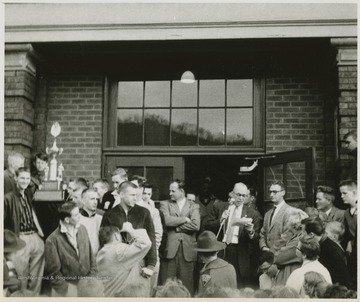A crowd celebrates the victory in front of the East Bank High School building. Jerry West is pictured in a striped shirt, standing beside a boy holding the trophy on the left of the photograph.West led the East Bank High School basketball team to its first ever West Virginia state championship title. He was named All-State from 1953–56, then All-American in 1956 when he was West Virginia Player of the Year, becoming the state's first high-school player to score more than 900 points in a season.West was born in Cheylan, W. Va. in 1938. After high school, he went on to play basketball for West Virginia University and then rose to fame as a player for the Los Angeles Lakers of the NBA before becoming a basketball coach and manager. 