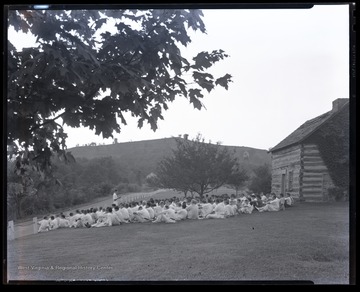 A group of campers sit on a lawn outside of a log cabin while listening to a speaker. 