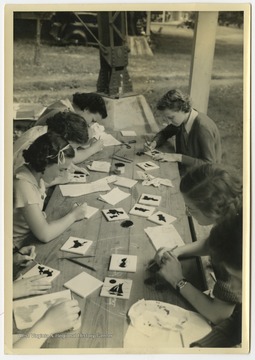 A group of unidentified girls paint together on a wooden table. 