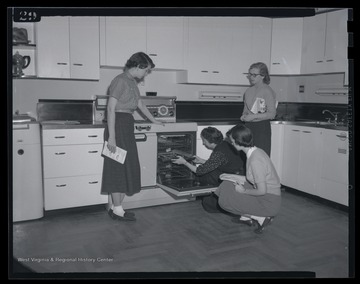 Three girls watch as their instructor inserts a pan into an oven. Subjects unidentified. 