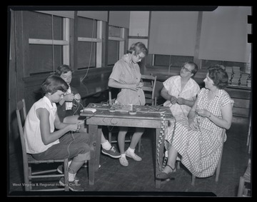 A girl tries on a hand-made belt while her companions weave threads together. Subjects unidentified. 