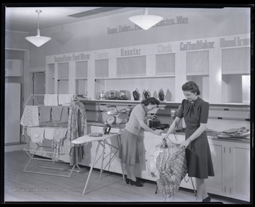 Two unidentified women are pictured ironing inside an exhibit of small electric household appliances. 
