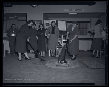 A group of women observe while two other women demonstrate how to clean carpets using an electric vacuum.