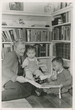 Pearl S. Buck sits on the floor with two small children and several books.  Caption on reverse reads "Pearl Buck with two Welcome House children".  The Welcome House was an adoption agency founded by Buck in 1949.