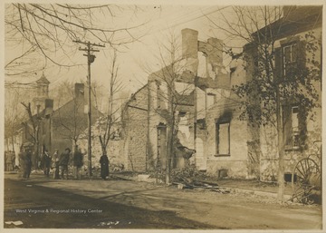 Rubble of several buildings after a fire, possibly in Berkeley or Jefferson County.  Several people stand in the street while one man looks into the ruins of a building.  There is a hose cart, used to fight fires, in front of one of the remaining buildings.