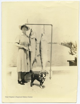 Louise holds onto her dog as she poses beside two large fish hanging from hooks. 