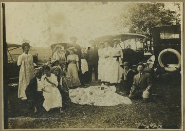 A group of men and women gather around to dine picnic-style. Around them are parked old-fashioned automobiles. Subjects unidentified. The photograph comes from a photo album belonging to Mrs. Earl R. Zinn.