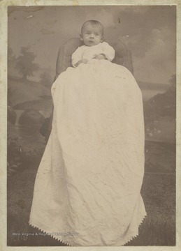 Infant Stella is dressed in a long white gown. 