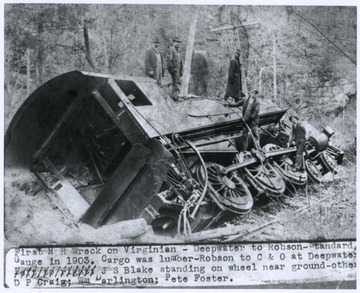 The train was carrying lumber from Robson, W. Va. to a C. & O. station at Deepwater, W. Va before wrecking. J. S. Blake is standing on the wheel near the ground. Also pictured are D. P. Craig, William Darlington, and Pete Foster. 