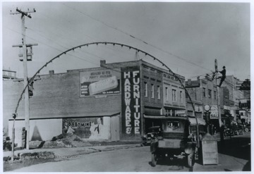 A number of automobiles are parked along the street. Visible signs include 'Hardware & Furniture,' an advertisement for Wrigley's gum, an advertisement for a play or movie called 'Baby Mine,' Ford and Chevrolet, and Frigidaire.