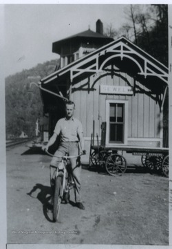 Omer Plumley is pictured riding a bicycle in the forefront, In the back is the Sewell Chesapeake and Ohio Railroad station. 