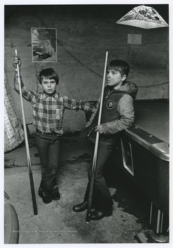The two boys are holding billiard sticks and are leaning against the pool table. 