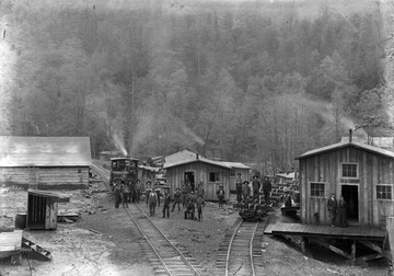 A group of mean are scattered across the rail tracks. On the left is a train engine. On the right appears to be a long rail cart designed to transport logs. 