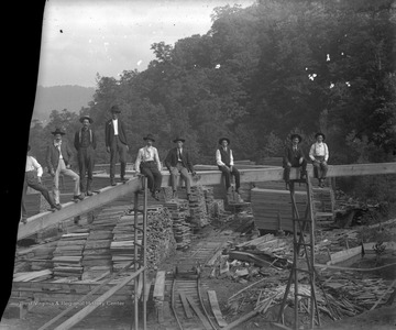 Lumbermen sit on top of a tall, wooden structure. In the background are piles of lumber. 