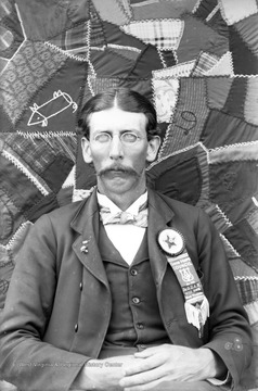 An unidentified man sits in front of a hanging quilt. The ribbon fastened to his jacket collar indicates he is the 153rd Good Hope Council member for the Junior Order of the United American Mechanics, an American fraternal order. It began as a youth affiliation of the Order of United American Mechanics, but seceded to become its own organization and eventually absorbed its parent order. Originally, it was an Anti-Catholic, Nativist group, but eventually abandoned this position and became a general fraternal benefit society open to people regardless of creed, race or sex.