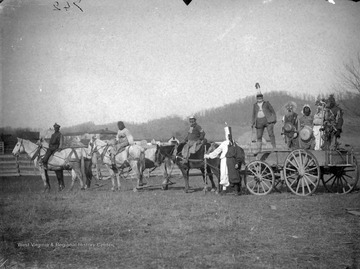 Nine unidentified individuals pose in costume. Three of them sit on horses. Five of them  stand on a carriage drawn by those horses, two of which wear large pots against their bellies. The man in the forefront, dressed in two, vertical striped colors, holds what appears to be a jousting rod.