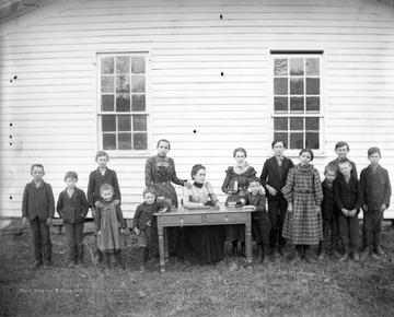A group of school children stand outside of the one-room school house. A woman, presumably the teacher, sits at a desk in the middle of the group.  The school was located on an old farm property belonging to the Post family.