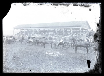 Horse-drawn vehicles are parked outside of the race track. In the background, a crowd fills the stands overlooking the track. 
