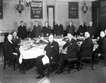 Executives of the Chevrolet Company gather around a table. In the background are Chevrolet advertisements. 
