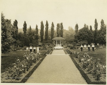 Eight men are pictured on either side of the lawn. The photograph looks down a pathway lined by flowers and directly at a gazebo. The garden is likely part of the property at Linden Hall in Pennsylvania.