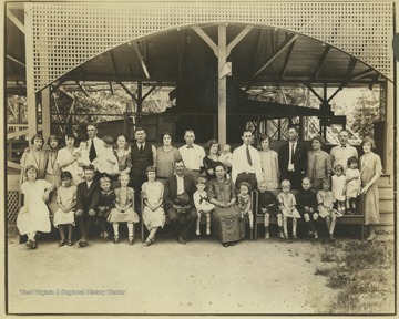 Men, women, and children pose for a group photo outside of a pavilion that leads to a roller coaster. 