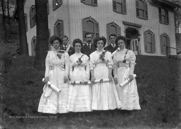 Four women stand in front of two boys and an older man, perhaps their professor. Each student holds a diploma.