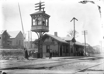A man, likely a railroad employee, stands in the forefront of the station by the tracks. In the background, groups of people lean against the building, perhaps waiting for a train.  In the foreground is the switch tower.