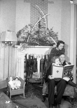 Young Patty Hoffman sits on her father's lap while he reads her the story of "Christmas," a book which is illustrated by a nativity scene. Behind them, Mrs. Hoffman reads along. Several stockings hang from the fireplace. Two dolls sit on a chair to the left. 