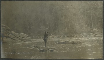 A man identified as "Dobbie" stands on top of a raft as he makes his way across the river.This photograph is found in a scrapbook documenting the survey for the Baltimore and Ohio Railroad in West Virginia and surrounding states. 