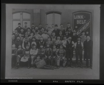 Group photo of men in front of Link-Belt Machinery sign.  One man holds a sign which identifies the group as the Materials Department.
