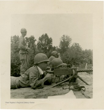 Man in prone position aiming machine gun down-range with other National Guard members in background.