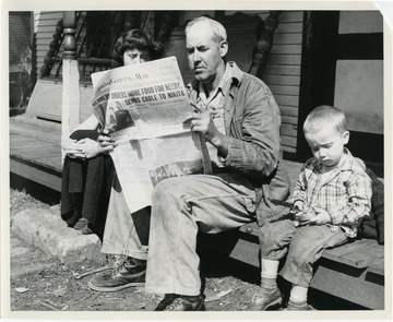 "Bud McDonald, one of the many persons now unemployed by the decrease in coal production in the area, reads a newspaper announcing President Kennedy's Executive Order Number 1, which made abundant agricultural commodities available to agencies for welfare distribution in areas of pressing need around the country, particularly those of high unemployment. Newspaper is dated January 22, 1961. With McDonald are Zelma Farmer and Jamie Deskin, both of Ethel, W. Va."