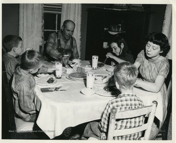 "A "Family" composed of members of community to represent typical family in this area, is shown eating a meal prepared completely from food distributed under President Kennedy's Executive Order Number 1. Clockwise, starting with man are: Bud McDonald; Patty Farmer; her mother, Mrs. Farmer; Billy Privet; and Jimmy and Richard Love."