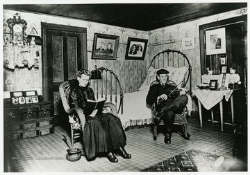 A woman sits reading a book and a man plays a fiddle cradled in the crook of his arm. 