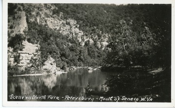 A view of the North Fork South Branch Potomac River at the spot Seneca Creek empties into it at the base of Seneca Rocks. In the foreground a man sits in a canoe.