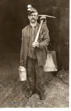 A coal miner stands with his pickaxe, shovel, and canister.