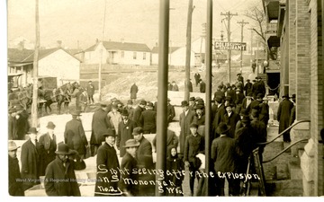 Crowds of men and women, with horse-drawn carriages, stand in the streets of Monongah. A sign for Coliseum Restaurant is visible in the background. Front of the card reads: ""Sightseeing after the explosion in S Monongah, W.Va."